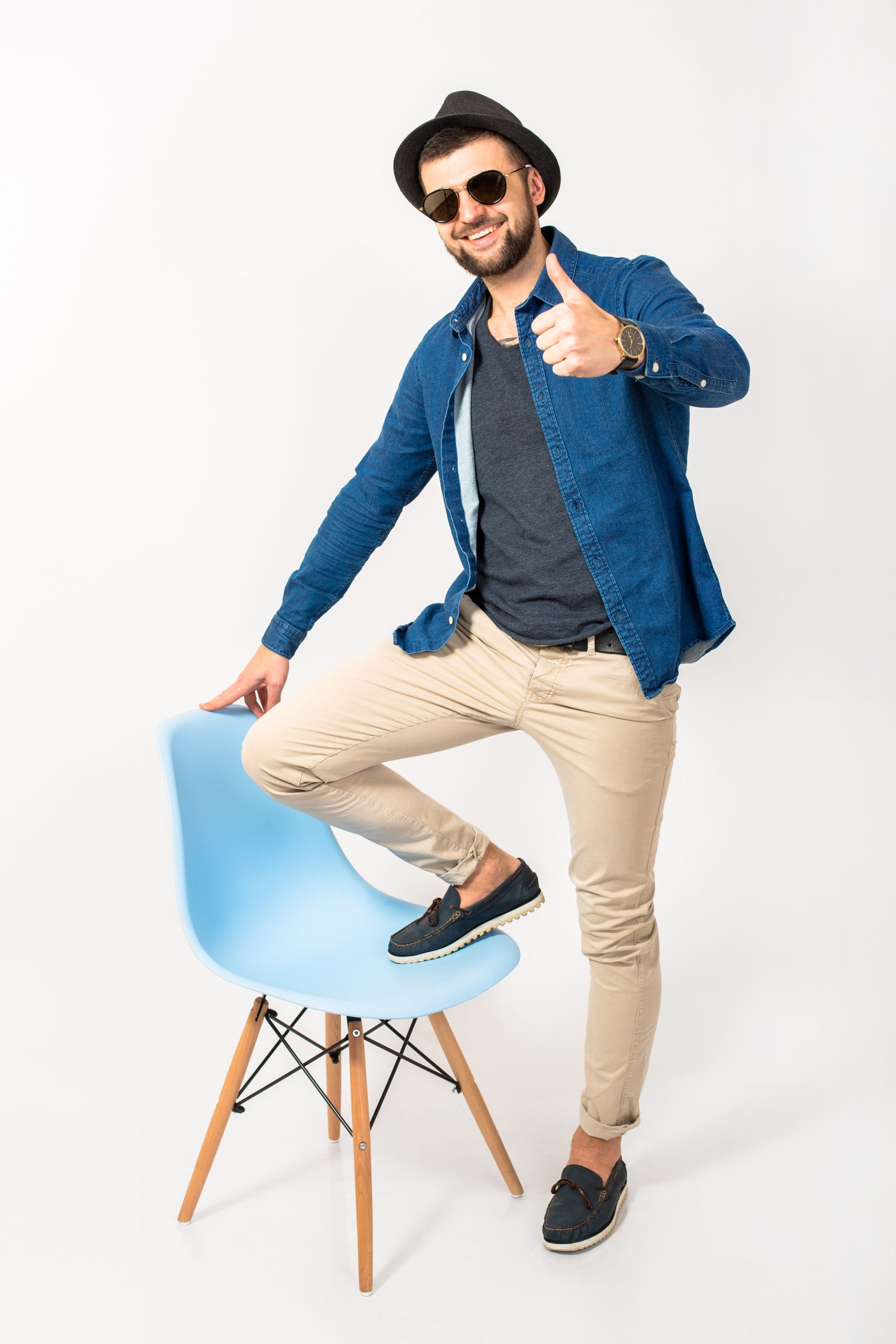 young handsome hipster man, isolated white studio background, stylish outfit, denim shirt, trousers, hat, sunglasses, standing on chair, cheerful, happy, smiling, positive, success, cheerful, thumb up